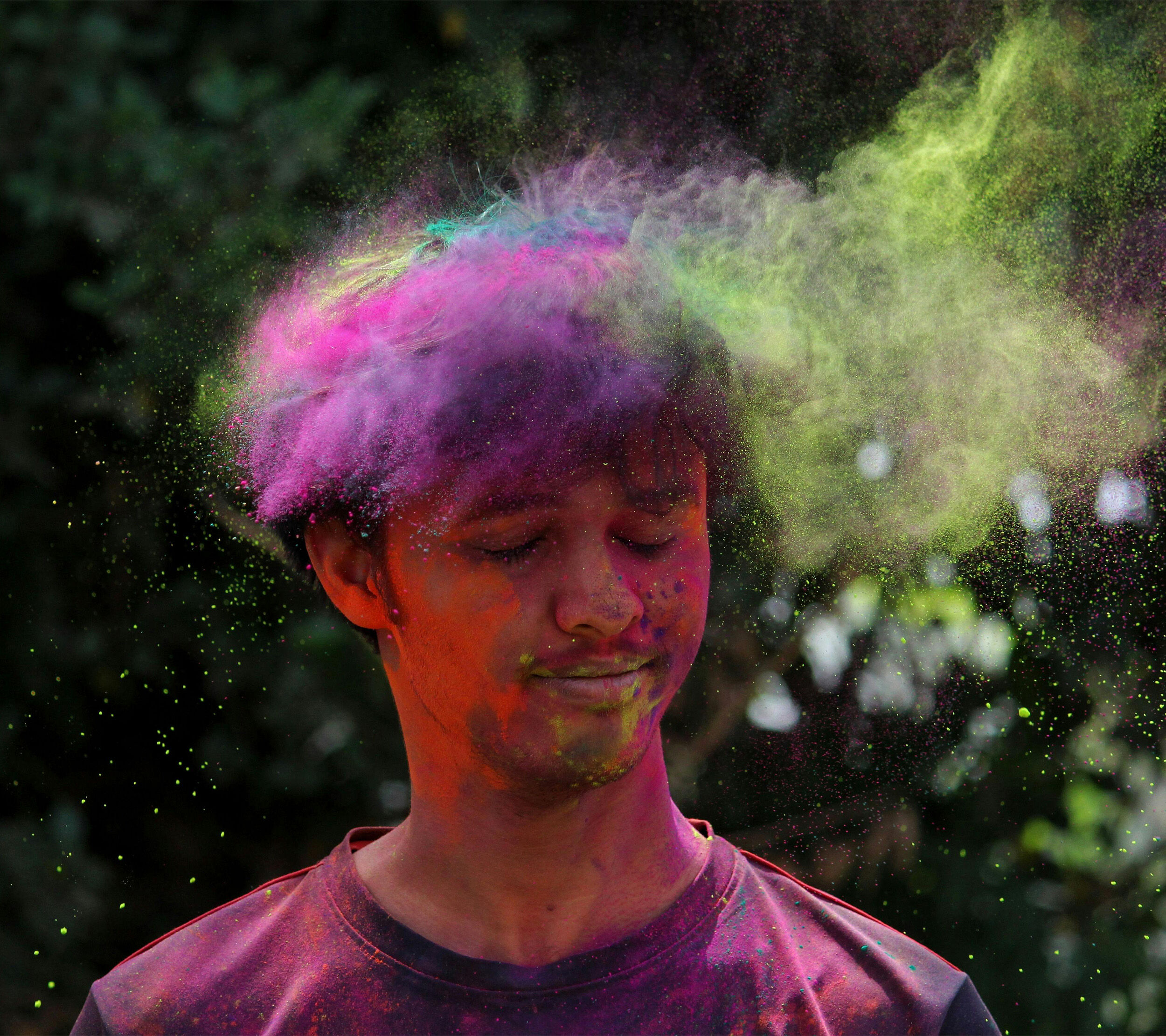 Close-up of a man outdoors as vibrant Holi powder clouds in pink, purple, and green sweep across his face and hair.